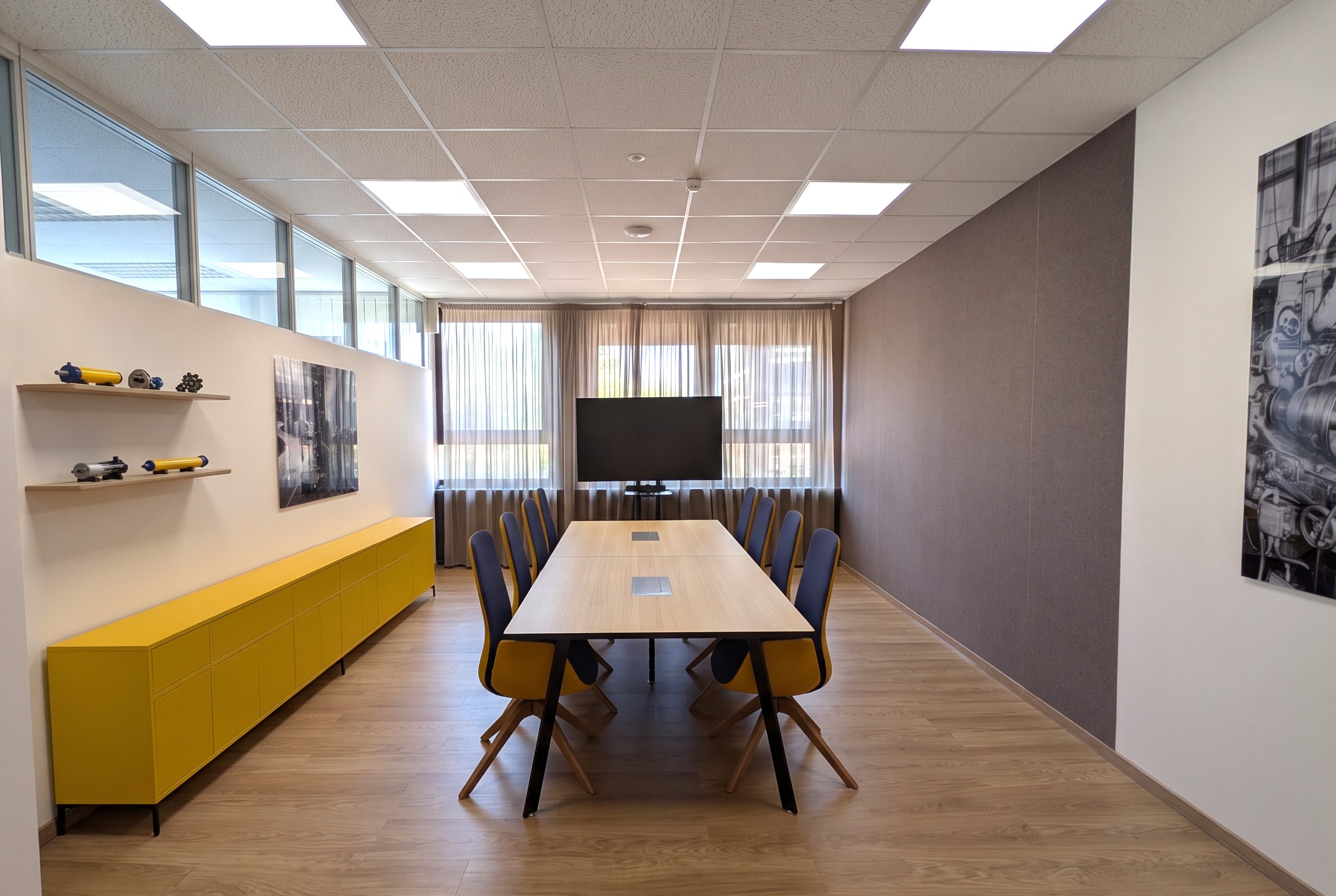 Zentrale Perspektive eines modernen Konferenzraums mit einem langen, mittig ausgerichteten Besprechungstisch und blau-gelben Polstersesseln. Eine dunkelgraue Akustikwand rechts balanciert das leuchtend gelbe Sideboard auf der linken Seite aus, während die quadratische Rasterdecke mit LED-Panels und die Oberlichter für eine gleichmäßige, helle Raumausleuchtung sorgen