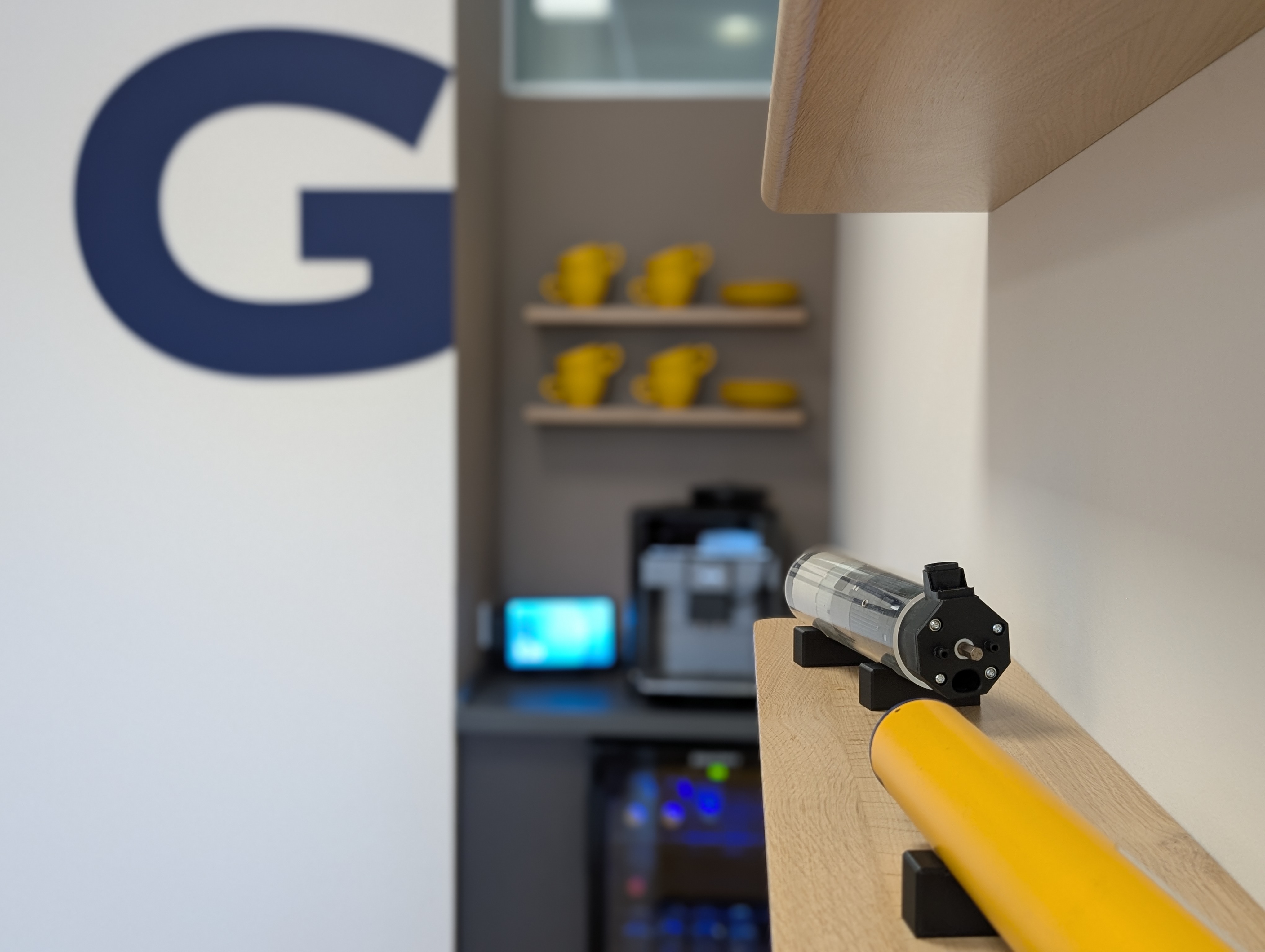 Detailaufnahme einer Büro-Kaffeestation mit Blick an schwebenden Holzregalen im Vordergrund vorbei. Im unscharfen Hintergrund sind ein Kaffeesurround, gelbe Tassen auf Regalen und ein blaues „G“-Wandbranding zu sehen, die das durchgängige Corporate-Color-Konzept des Workspace-Designs unterstreichen
