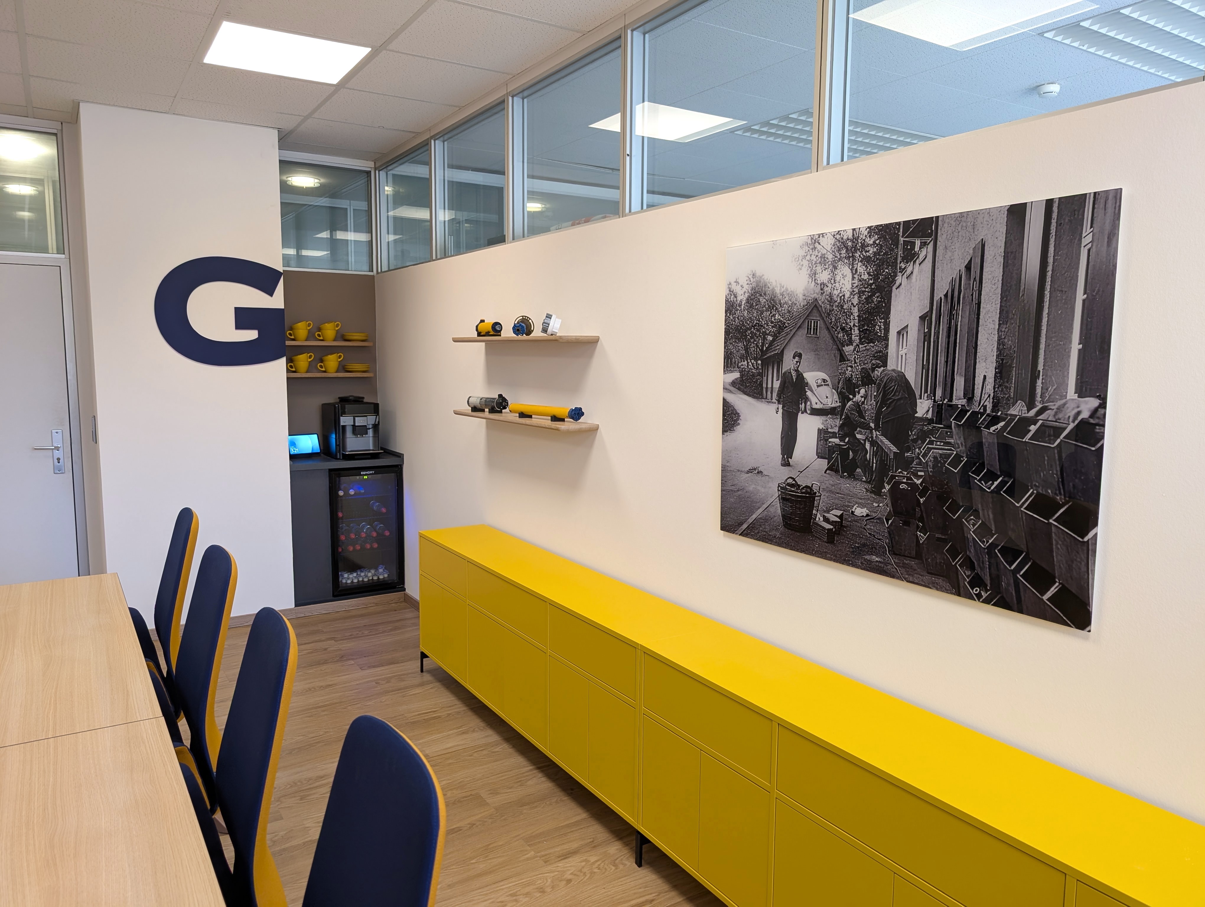 Perspektivische Ansicht eines Konferenzraums mit einem langen, leuchtend gelben Sideboard an einer weißen Wand. Ein großformatiges Schwarz-Weiß-Bild, schwebende Holzregale und ein blaues Firmenlogo rahmen die Sichtachse zur Kaffeestation im Hintergrund, während links die Rückenlehnen der blau-gelben Konferenzstühle in Reihe angeordnet sind