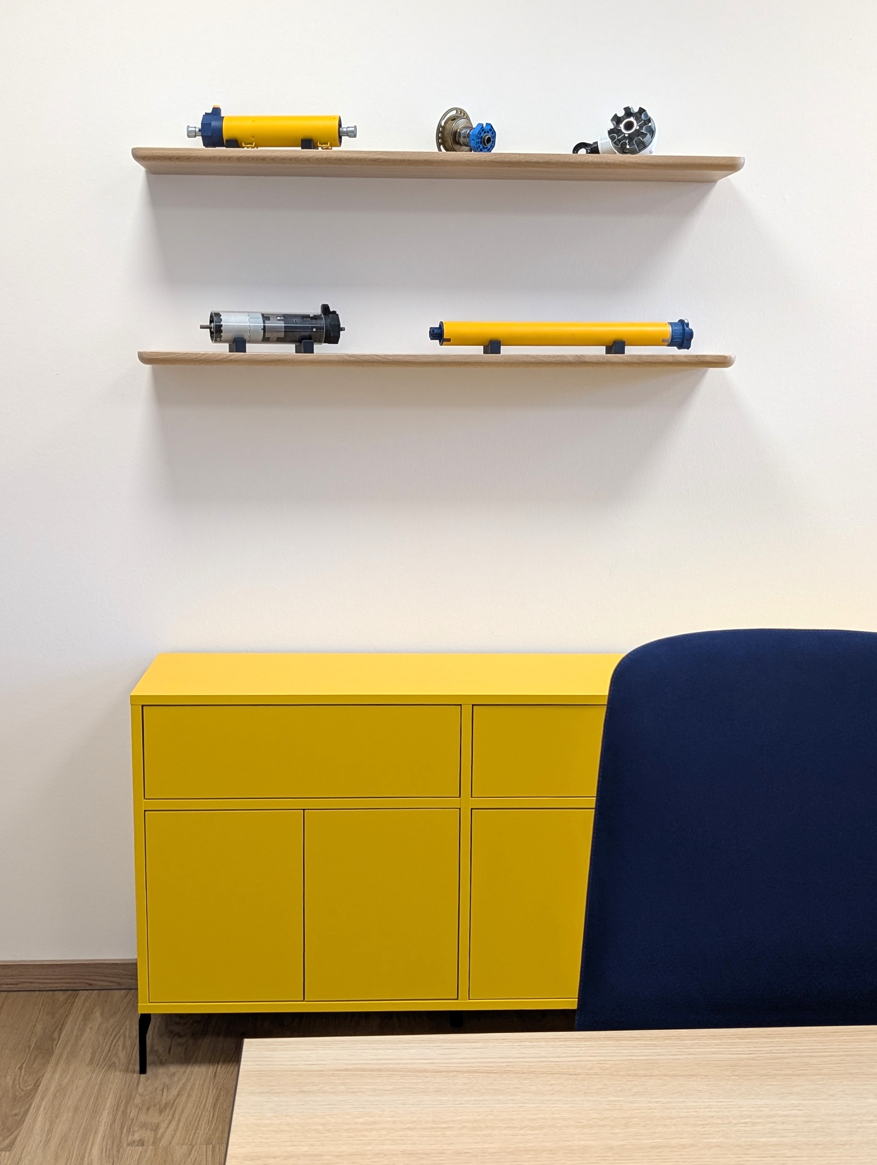 Detailansicht einer Büroeinrichtung mit einem grifflosen Sideboard in markantem Postgelb auf filigranen schwarzen Metallfüßen. Darüber befinden sich zwei schwebende Holzregale aus heller Eiche, auf denen technische Exponate präsentiert werden, während im rechten Vordergrund eine dunkelblaue Stuhlrückenlehne und eine Tischkante die Raumperspektive rahmen