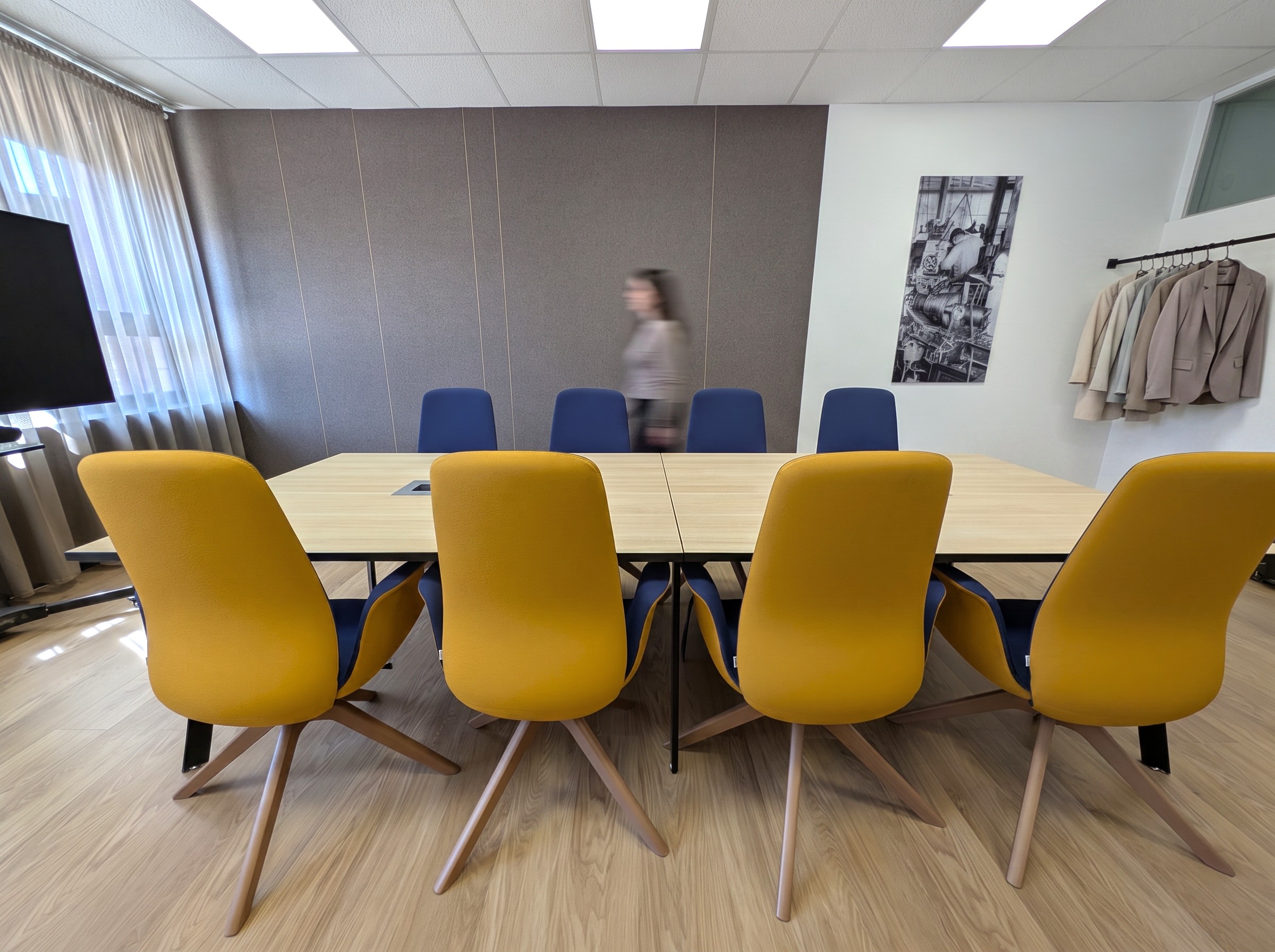 Moderner Konferenzraum mit einem langen Besprechungstisch in heller Holzoptik und einem kontrastierenden Bestuhlungskonzept aus senfgelben Polstersesseln im Vordergrund und blauen Stühlen im Hintergrund. Eine dunkelgraue Akustik-Paneelwand, ein heller Vinylboden und eine Person in Bewegungsunschärfe unterstreichen die funktionale Arbeitsatmosphäre