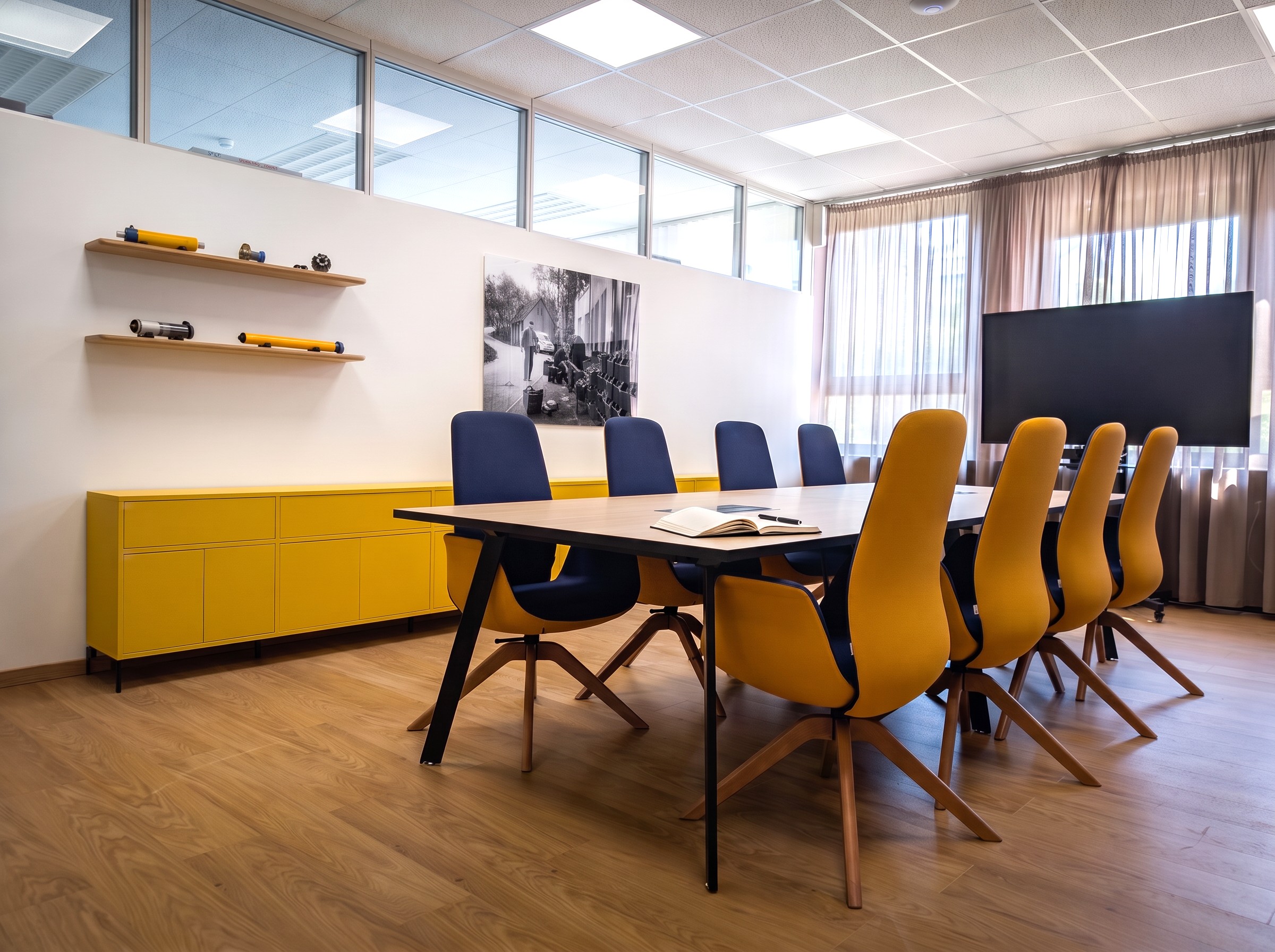 Moderner Konferenzraum mit einem großen Besprechungstisch, umgeben von senfgelben und dunkelblauen Polsterstühlen auf einem hellen Holzboden. Das Raumkonzept integriert ein leuchtend gelbes Sideboard als farbigen Akzent, schwebende Holzregale an einer weißen Trennwand mit Oberlichtern sowie einen großen Präsentationsbildschirm vor einer transparenten Fensterfront mit Vorhängen