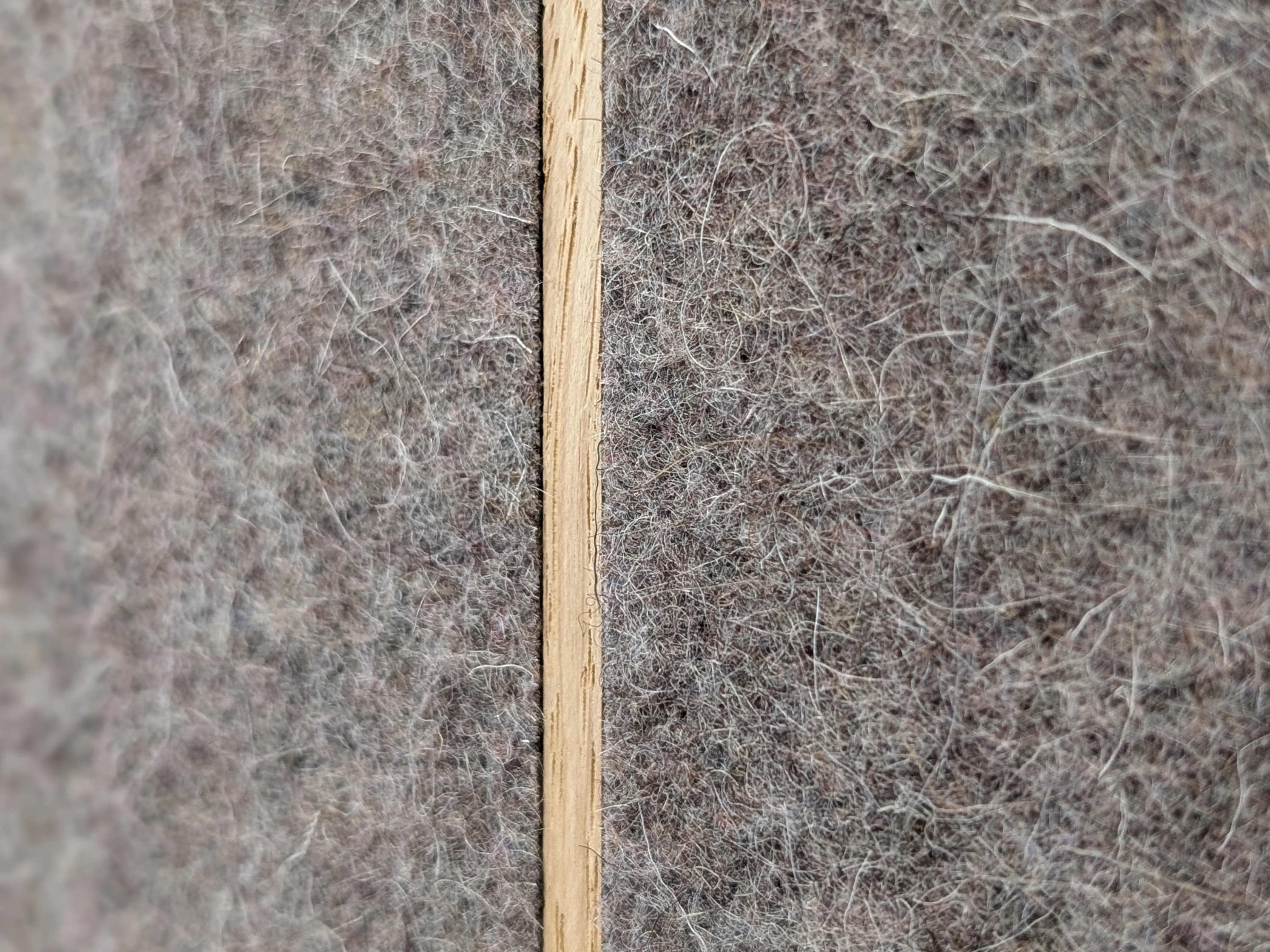 Makroaufnahme eines Akustikpaneels mit einer strukturierten Oberfläche aus grauem, meliertem Filzstoff und einer vertikal verlaufenden Holzlatte aus heller Eiche. Die detailreiche Ansicht betont die weiche, faserige Textur des schallschluckenden Trägermaterials im Kontrast zur glatten, natürlichen Holzmaserung